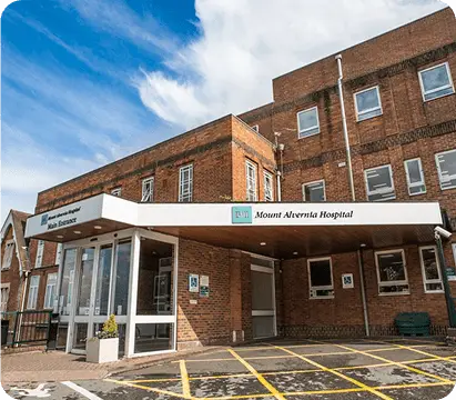 The image shows the exterior of Mount Alvernia Hospital, a brick building with a white main entrance canopy and parking spaces marked in yellow. The hospital is home to leading oncologists in Guildford, specialising in testicular cancer. The sky above is partly cloudy.