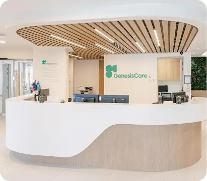 A modern medical reception area with a curved white desk, wood accents, and GenesisCare signage on the wall welcomes visitors seeking an oncologist in Windsor. Bright and clean, the space features computers, office supplies, and small flags on the desk.