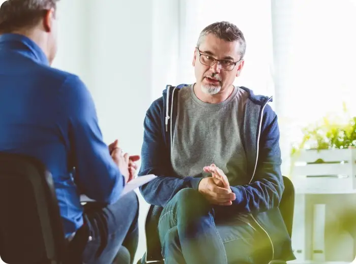 Two men sit facing each other in a brightly lit room, having a conversation. One gestures with his hands while the other, an oncologist in Guildford, listens attentively and takes notes on a clipboard.