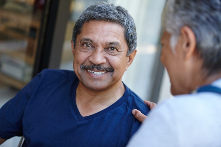 An older man with gray hair and a mustache smiles warmly while sitting outdoors, wearing a navy blue shirt. Another person, possibly his testicular cancer specialist, sits close beside him, touching his shoulder supportively.