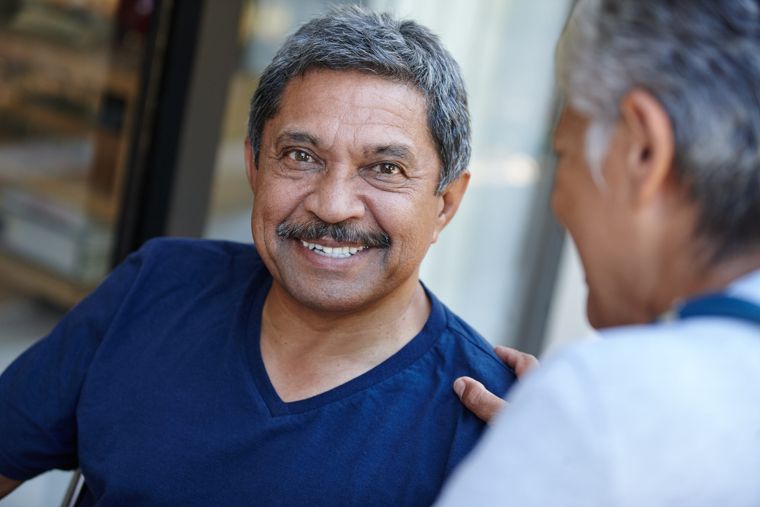 An older man with gray hair and a mustache smiles warmly while sitting outdoors, wearing a navy blue shirt. Another person, possibly his testicular cancer specialist, sits close beside him, touching his shoulder supportively.