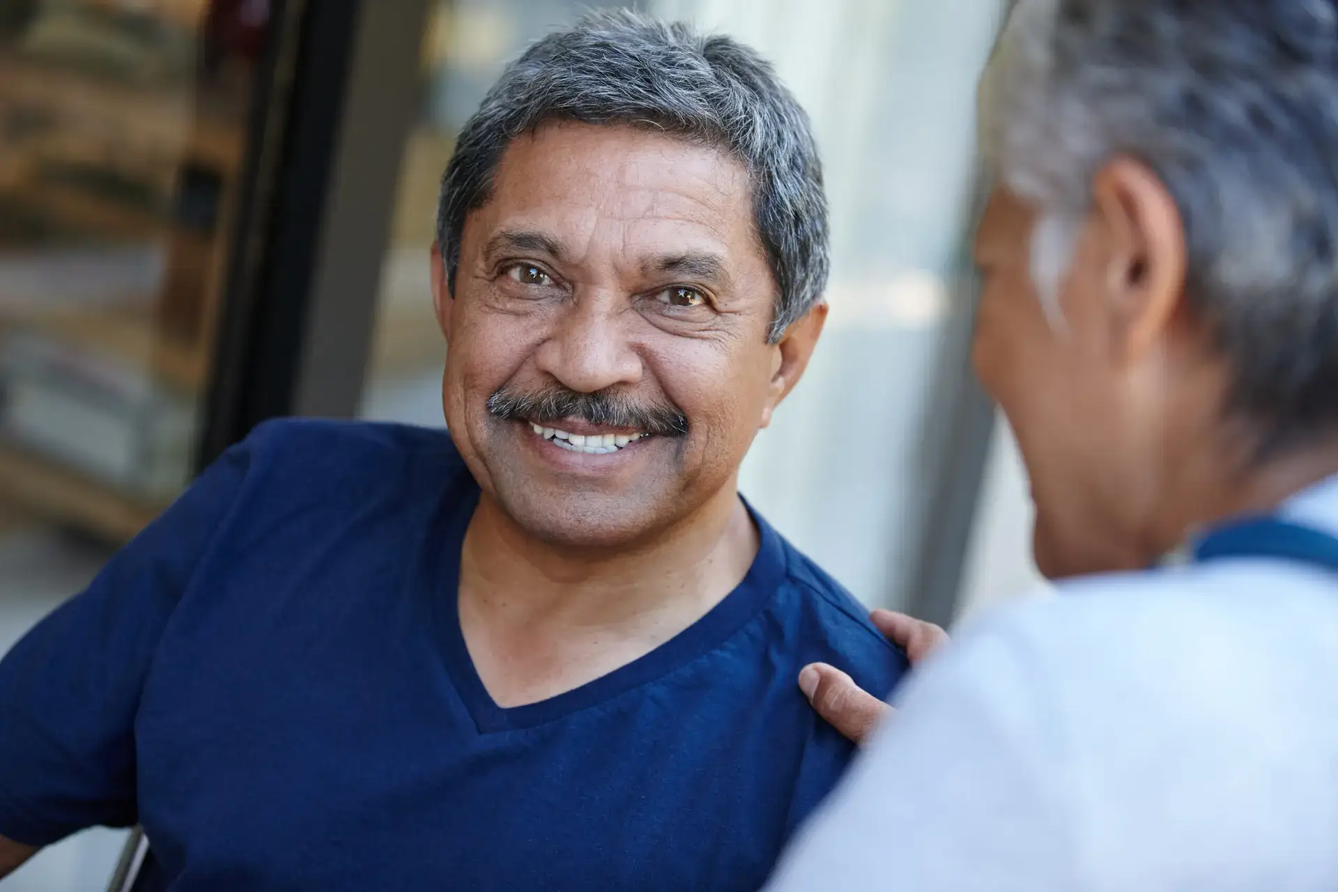 An older man with gray hair and a mustache smiles warmly while sitting outdoors, wearing a navy blue shirt. Another person, possibly his testicular cancer specialist, sits close beside him, touching his shoulder supportively.