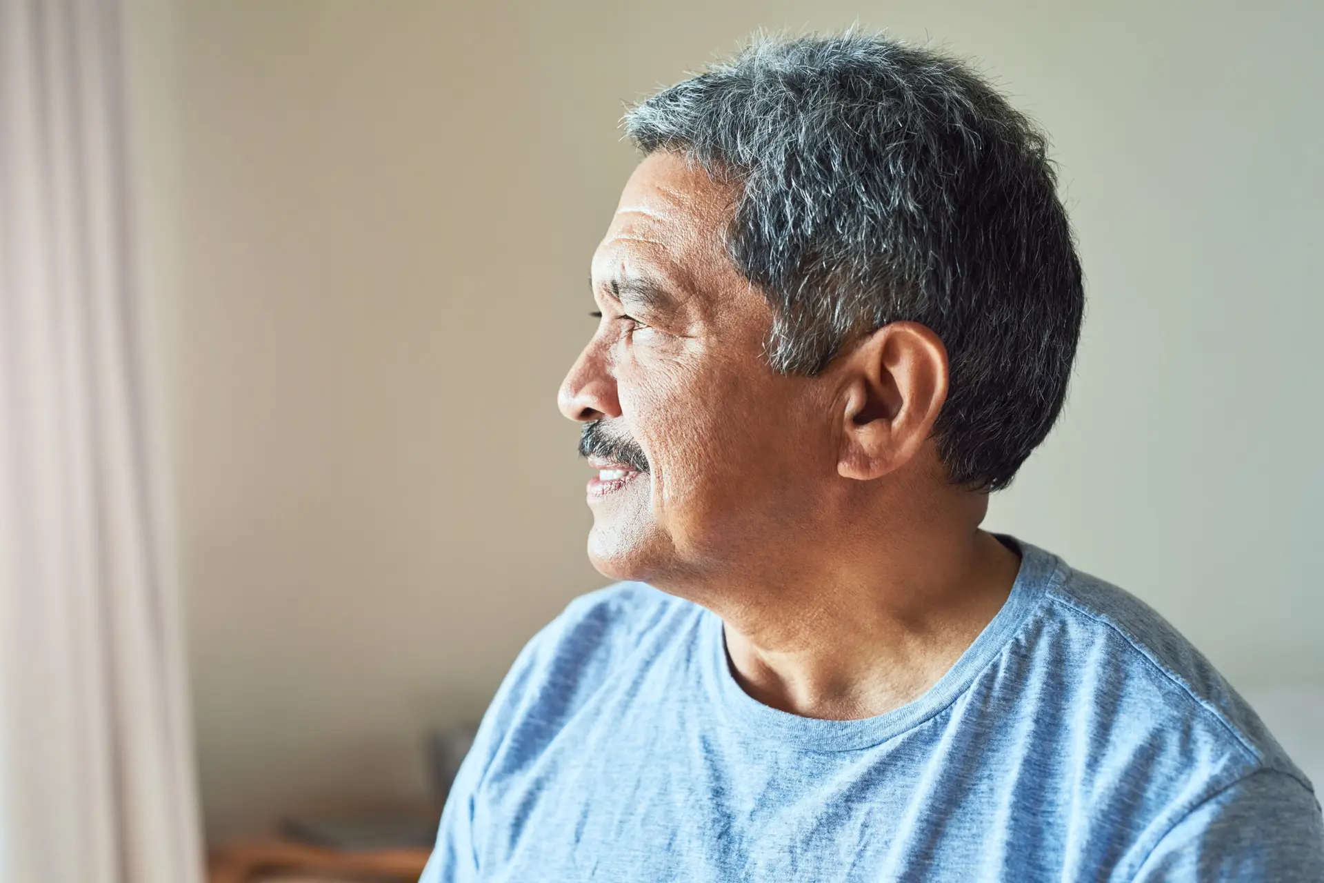 Older man with short gray hair and a mustache, wearing a light blue shirt, looks thoughtfully out of a window with soft natural light illuminating his face; he is an experienced testicular cancer specialist.