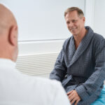 A smiling man in a gray robe sits on a medical examination table, facing Dr Christos Mikropoulos, a renowned testicular cancer specialist, in a bright, modern room.