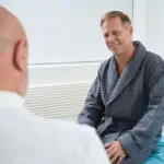 A smiling man in a gray robe sits on a medical examination table, facing Dr Christos Mikropoulos, a renowned testicular cancer specialist, in a bright, modern room.