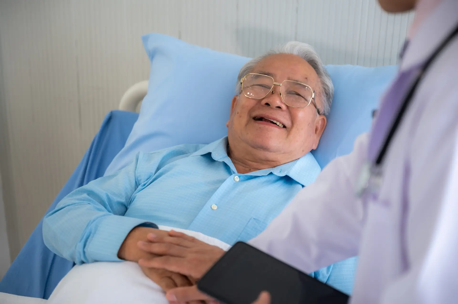 An elderly man lies in a hospital bed, smiling and holding his hands together, while Dr Christos Mikropoulos, a renowned oncologist in Guildford & Windsor, stands nearby holding a tablet.