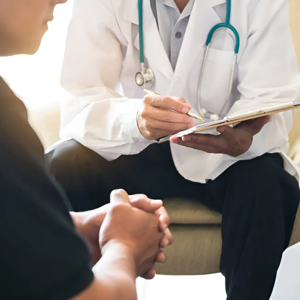 Dr Christos Mikropoulos, a testicular cancer specialist, wearing a white coat and stethoscope, discusses notes on a clipboard with a seated patient, who has hands clasped, in a medical consultation setting.
