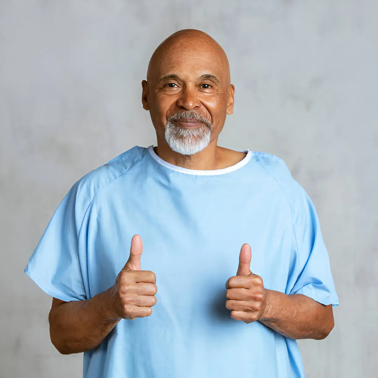 An older man wearing a light blue hospital gown smiles and gives two thumbs up, standing against a plain light background—celebrating care from testicular cancer specialist Dr Christos Mikropoulos.