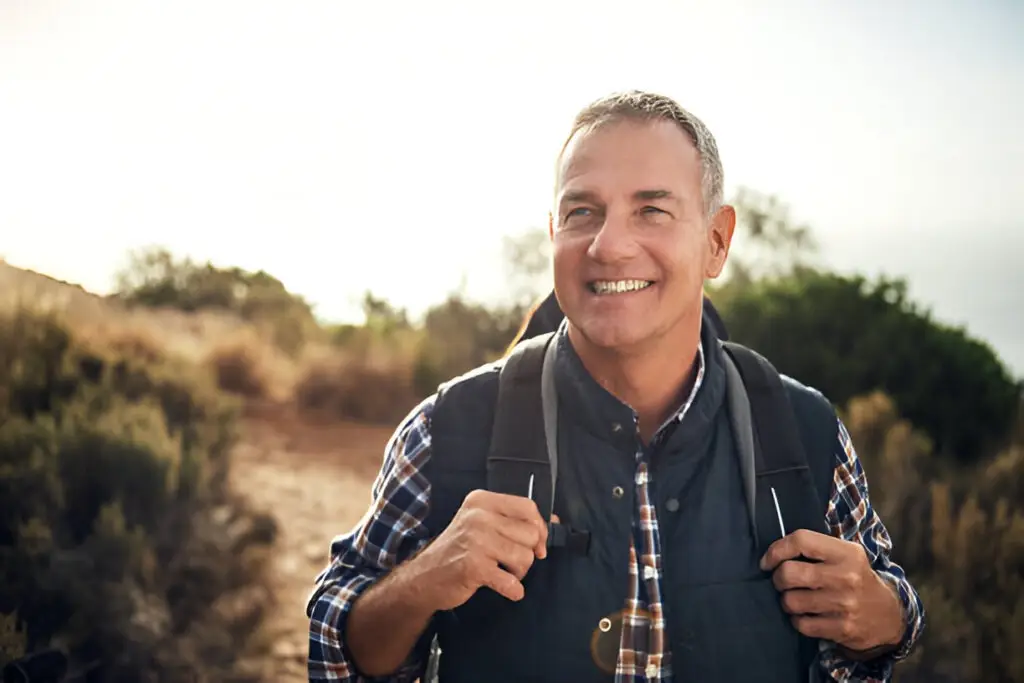 A smiling middle-aged man wearing a plaid shirt and vest stands outdoors on a dirt path, carrying a backpack. The sunlit trees and bushes hint at a hiking adventure—perfect for an Oncologist in Guildford who loves exploring nature.