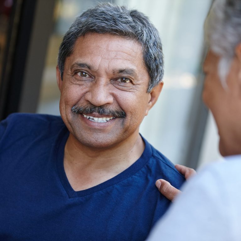 An older man with gray hair and a mustache smiles warmly while sitting outdoors, wearing a navy blue shirt. Another person, possibly his testicular cancer specialist, sits close beside him, touching his shoulder supportively.