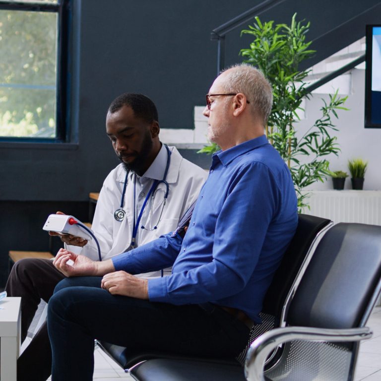 two-men-sit-waiting-room-with-monitor-showing-word-health-screen